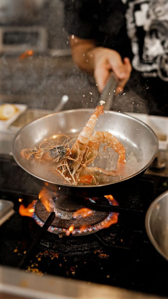 A chef expertly cooks prawns in a frying pan over a gas flame in a restaurant kitchen.